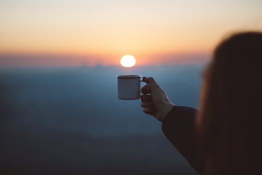 A person holds a coffee cup against a beautiful sunrise, capturing a serene outdoor moment.